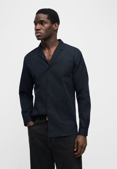 Νεαρός με κοντά σγουρά μαλλιά, φορώντας σκούρο μπλε πουκάμισο με μακριά μανίκια και μαύρο τζιν, στέκεται με το ένα χέρι στην τσέπη.