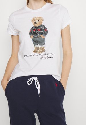 Camiseta blanca con un oso de peluche que lleva un suéter estampado, pantalones y botas, combinada con pantalones azul marino con cordón y un logo rojo bordado.