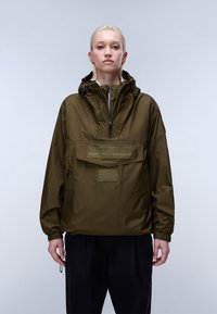 Νεαρό άτομο με κοντά ξανθά μαλλιά, που φοράει μια ελιάς πράσινη ζακέτα με κουκούλα και μαύρο παντελόνι, στέκεται μπροστά σε απλό ανοιχτόχρωμο φόντο.