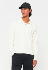 Langermet polo skjorte i off-white stoff med krage og knepping, med rett kant og brukt sammen med mørke bukser og en svart caps.