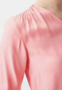 Rosa Bluse mit V-Ausschnitt, mit gefälteltem Detail an der Schulter und einer glatten, satinähnlichen Textur. Halb-taillierter Schnitt.