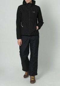 Persona che indossa una giacca a cappuccio nera e pantaloni neri, in piedi con una mano in tasca e stivali da trekking marroni con lacci gialli.