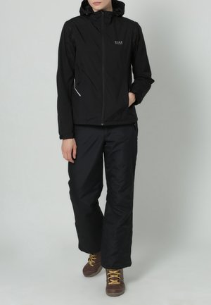 Persona vestida con chaqueta con capucha negra y pantalones negros, de pie con una mano en el bolsillo y botas de senderismo marrones con cordones amarillos.