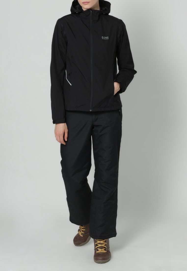 Persona che indossa una giacca a cappuccio nera e pantaloni neri, in piedi con una mano in tasca e stivali da trekking marroni con lacci gialli.