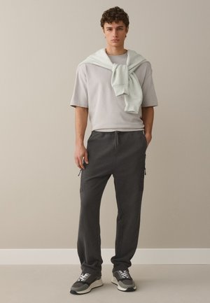 Joven de pie, vistiendo una camiseta gris clara, pantalones de chándal gris oscuro, zapatillas grises y una sudadera ligera sobre los hombros.