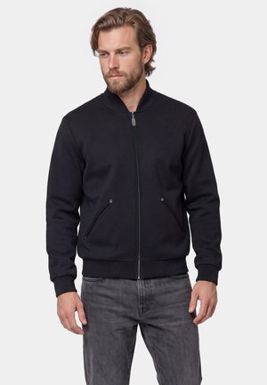 Man wearing a black zip-up bomber jacket with ribbed cuffs and collar, paired with gray jeans, looking to the side.
