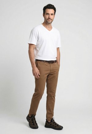 Man standing with hands in pockets wearing white V-neck T-shirt, brown pants, black belt, and black sneakers against plain background.