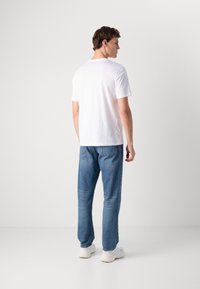 Mann som står med ryggen til, iført hvit t-skjorte, blå jeans og hvite sneakers mot en enkel lys bakgrunn.