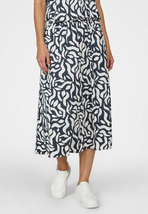 Midirock mit schwarz-weißem abstraktem Blattmuster, getragen mit weißen Turnschuhen, Person steht vor einfarbigem Hintergrund.