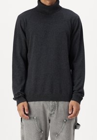 Uomo che indossa un maglione a collo alto grigio scuro e pantaloni larghi grigi chiari con grandi tasche frontali, in piedi contro uno sfondo bianco.