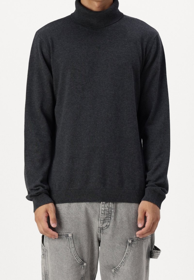 Uomo che indossa un maglione a collo alto grigio scuro e pantaloni larghi grigi chiari con grandi tasche frontali, in piedi contro uno sfondo bianco.