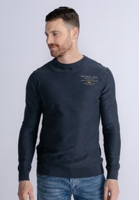 Granatowy bawełniany sweatshirt z długimi rękawami, okrągłym dekoltem i ściągaczami. Na piersi znajduje się logo w jasnym beżu.