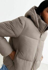 Pufferjack in licht taupe met een capuchon, geborduurd ontwerp en een ritssluiting aan de voorkant. Beschikt over elastische manchetten en zijzakken. Gladde textuur.