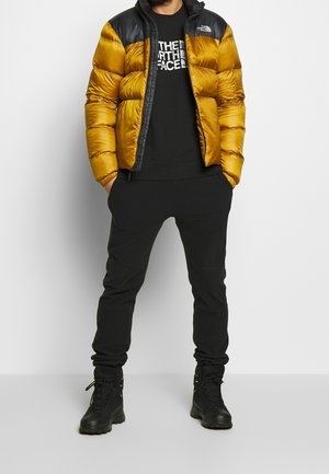 Persona con giacca imbottita giallo senape e nera, t-shirt nera The North Face, pantaloni neri e stivali neri con lacci, in piedi con le mani nelle tasche.