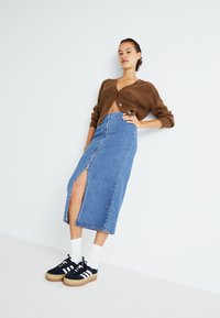 Rjava rebrasta kardigan, modra midi krilo iz denima z sprednjim razporkom, črnobeli superge na belih nogavicah. Minimalistična ozadje.