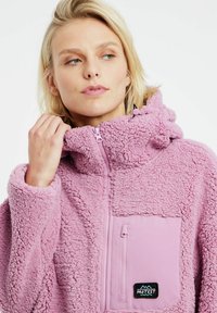 Giacca in pile rosa con texture morbida, apertura frontale a mezza zip, cappuccio e tasca sul petto con un'etichetta del marchio nera.