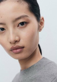 Gros plan sur une personne ayant la peau lisse, les yeux foncés et les cheveux noirs attachés, portant un haut gris devant un fond clair uni.