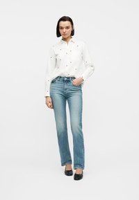 Donna in piedi che indossa una camicia bianca con piccoli cuori rossi, jeans blu a gamba dritta e ballerine nere, posando con una mano in tasca.