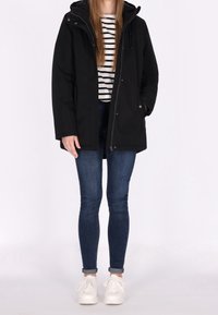 Zwarte parka met capuchon, ritssluiting en drukknopen, gecombineerd met een gestreept shirt en blauwe skinny jeans, afgewerkt met witte sneakers.