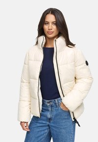 Giacca in crema imbottita con colletto alto e zip frontale, con un accento nero sulla manica. Abbinata a un top blu scuro e jeans a vita alta.
