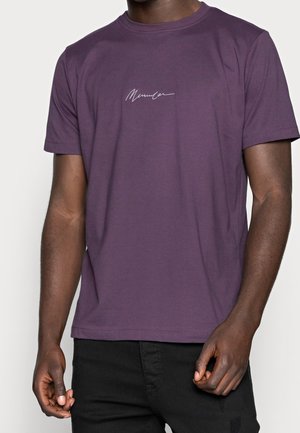 Homme portant un T-shirt violet à manches courtes avec un petit texte blanc de style signature centré sur la poitrine.
