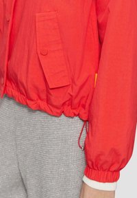 Raudona lengva striukė su trumpa iškirpte, elastingais rankogaliais, priekinė kišenė su spaustuku ir reguliuojamu juosmeniu. Dėvima ant tekstūruotų pilkų kelnių.