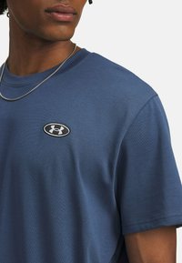 Short-sleeve, blue t-shirt made of soft fabric. Features a black and white oval logo on the chest. Simple design with a crew neckline.