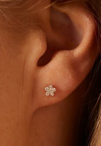Oreille avec une petite boucle d'oreille en forme de fleur composée de cinq gemmes claires sur le lobe de l'oreille.