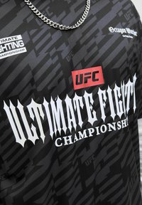 Chemise noire de l'UFC (Ultimate Fighting Championship) avec un texte gothique blanc, un logo UFC rouge, et une chaîne en argent autour du cou d'une personne.
