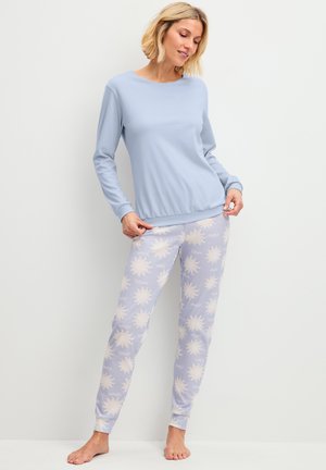 Vrouw draagt een lichtblauw longsleeve top en lichtpaarse pyjamabroek met crèmekleurige zonpatronen, staat op blote voeten tegen een witte muur.