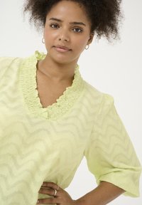 Blusa de color amarillo claro con un patrón de zigzag texturizado, cuello con volantes y mangas abullonadas. Confeccionada en una tela suave y ligera.