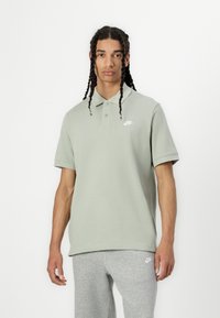 Green cotton polo shirt with short sleeves, three-button collar, and white Nike logo on the left chest, paired with gray sweatpants.