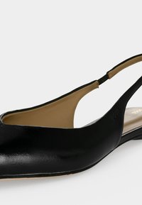 Chaussure plate à bride arrière en cuir noir avec un intérieur beige et un bout arrondi, présentée sur un fond gris clair uni.