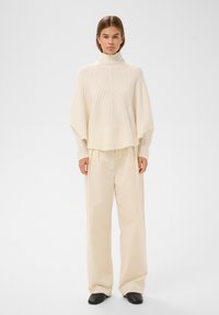 Kobieta w kremowym swetrze z golfem o prążkowanej fakturze z szerokimi rękawami, luźnych, wysokich spodniach w kremowym kolorze i czarnych płaskich butach, stojąca na białym tle.