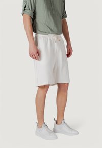 Una persona che indossa una camicia verde chiaro e pantaloni corti bianchi si trova, abbinata a sneakers bianche, su uno sfondo neutro.