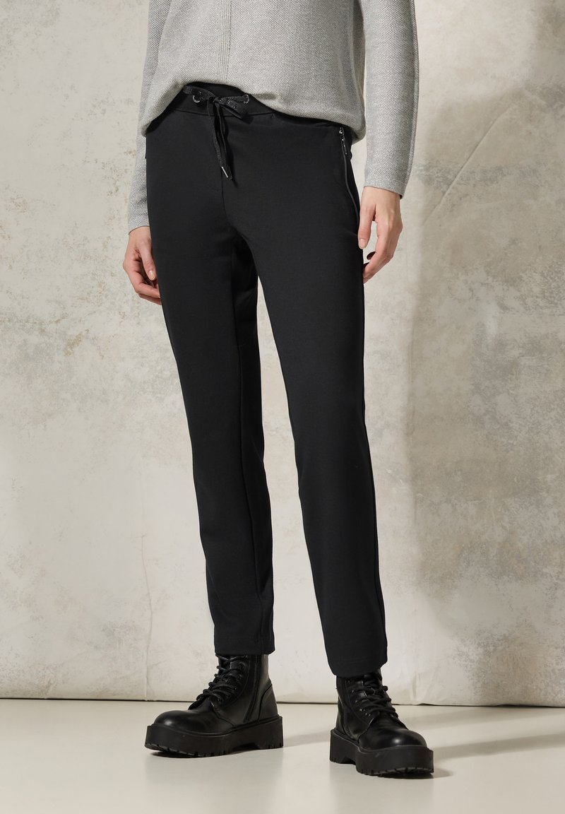 Black tailored pants with a slim fit, featuring a drawstring waistband and zippered side pockets. Worn with lace-up black boots.