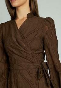 Rochie maro dintr-un material texturat, cu modele florale intricate, mâneci lungi și un detaliu de talie legat. Prezintă un guler înalt.