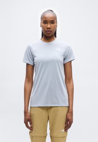 Γυναίκα που φοράει γκρι ανοιχτό κοντομάνικο πουκάμισο The North Face και μπεζ παντελόνι με αποσπωμένα πόδια, στέκεται μπροστά σε απλό λευκό φόντο.