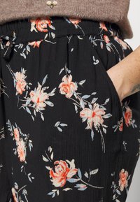 Pantalon en tissu noir avec un motif floral comprenant des roses pêche et rose clair, taille élastique, et main reposant à l'intérieur de la poche.