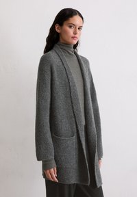Šedý pletený cardigan se strukturovaným povrchem, otevřeným předním dílem a dvěma kapsami. Dlouhé rukávy vrstvené přes přiléhavý rolák.