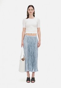 Nő fehér, rövidített, texturált felsőt, kék pliszírozott midi szoknyát, fekete nyitott orrú magassarkút, arany fülbevalót visel, miközben egy nagy fehér táska van a kezében.