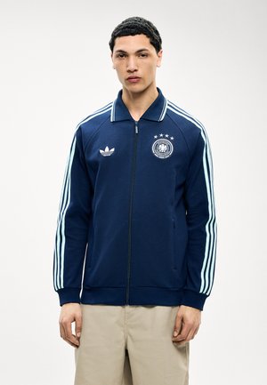 Noor mees, kel sinakas-musta jahedusega spordijakk, mille varrukatel on helesinised triibud ning rinnal Saksa jalgpallimeeskonna märk.