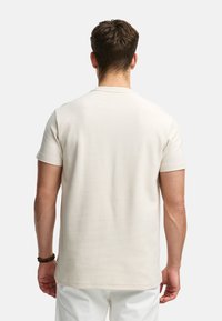 Camiseta de manga corta beige con un acabado texturizado. Diseño de cuello redondo, corte relajado, presenta detalles de costura sutiles, combinada con pantalones blancos.