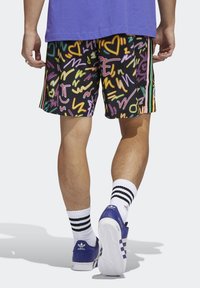 Shorts neri con motivi astratti colorati, con dettagli verdi, gialli, rosa e viola. Abbinati a calzini bianchi e scarpe blu.