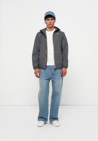 Chaqueta cortaviento gris con capucha, detalles en cremallera amarillos, llevada sobre una camiseta blanca. Jeans de pierna ancha azul claro y una gorra azul claro.
