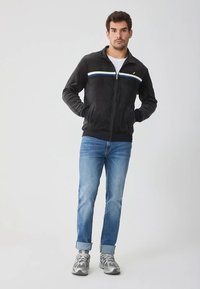 Giacca in pile nera con cerniera completa, strisce orizzontali bianche e blu, e un piccolo logo. Abbinata a jeans azzurri e sneakers grigie.