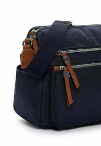 Navyfarbener Stoffbeutel mit braunen Lederakzenten, zwei vorderen Reißverschlüssen, einem breiten verstellbaren Schultergurt und einer strukturierten Oberfläche.