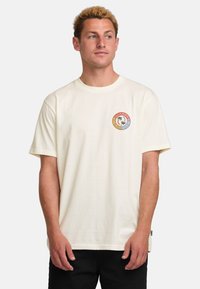 Cream-colored cotton t-shirt with short sleeves, round neckline, and a circular multicolor logo on the left chest featuring a stylized design.