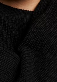 Svart stickad tröja med en texturerad, ribbad mönstring och en lös krage. Materialet verkar mjukt och varmt. Närbild.