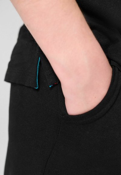 Main partiellement dans la poche avant d'un pantalon noir porté avec une chemise noire avec un détail d'ourlet turquoise.